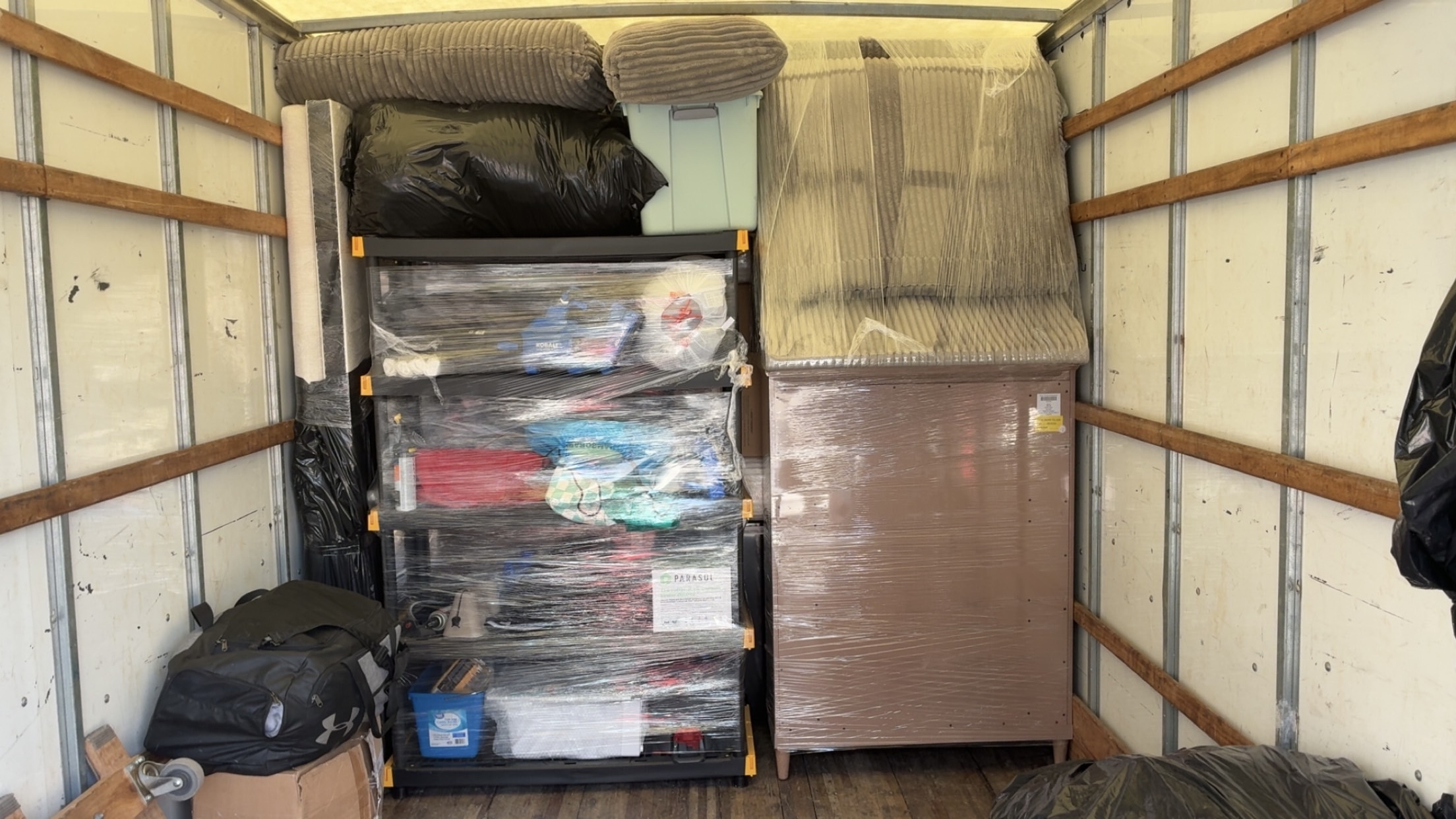 The interior of a loaded moving truck with a shrink-wrapped shelving unit, a wrapped wooden dresser, a plastic-covered mattress and sofa cushions packed tightly floor to ceiling