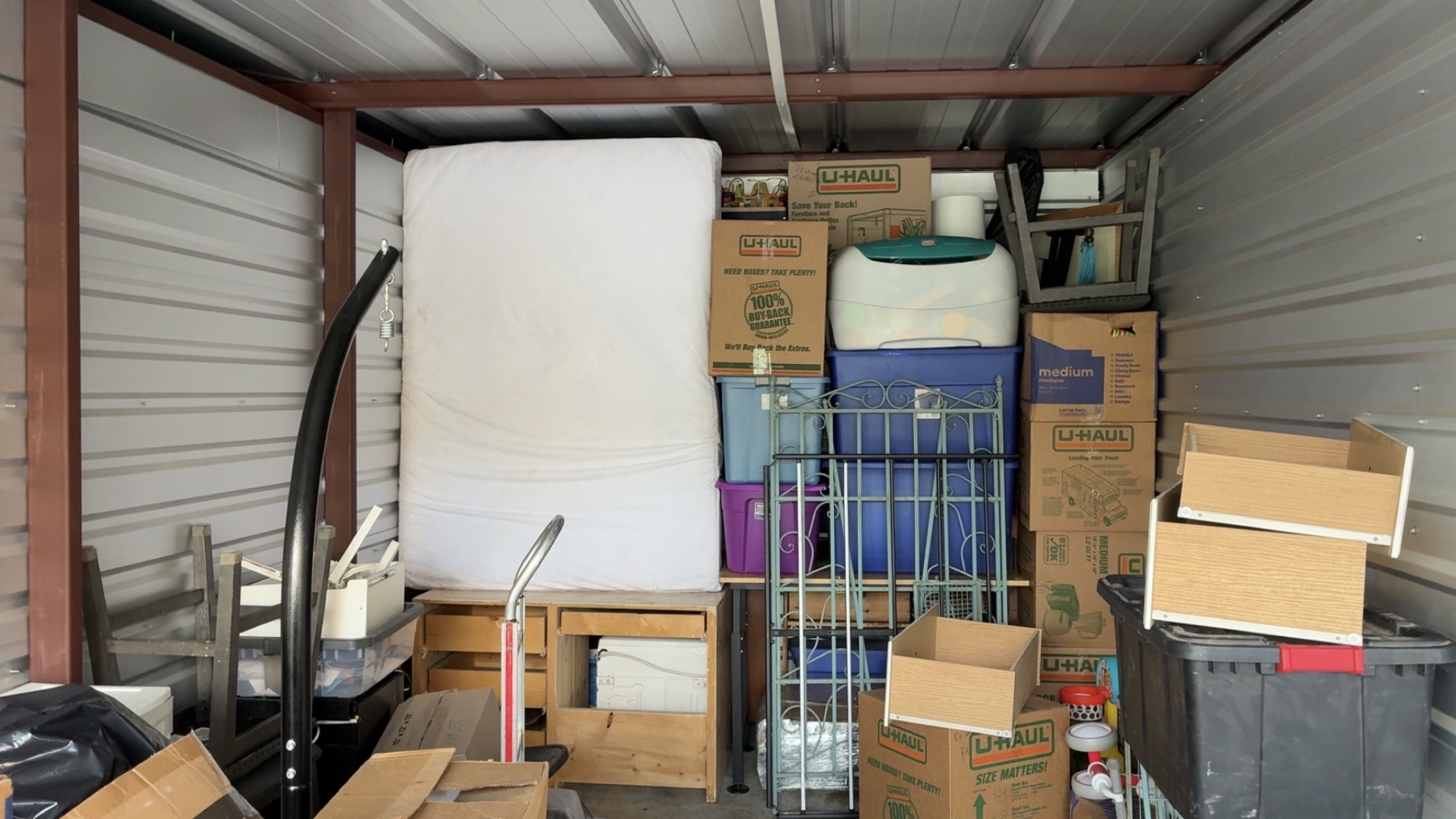 A neatly packed self-storage unit with an upright mattress, stacked labeled boxes, plastic totes, a metal garden trellis and wooden furniture organized against the walls